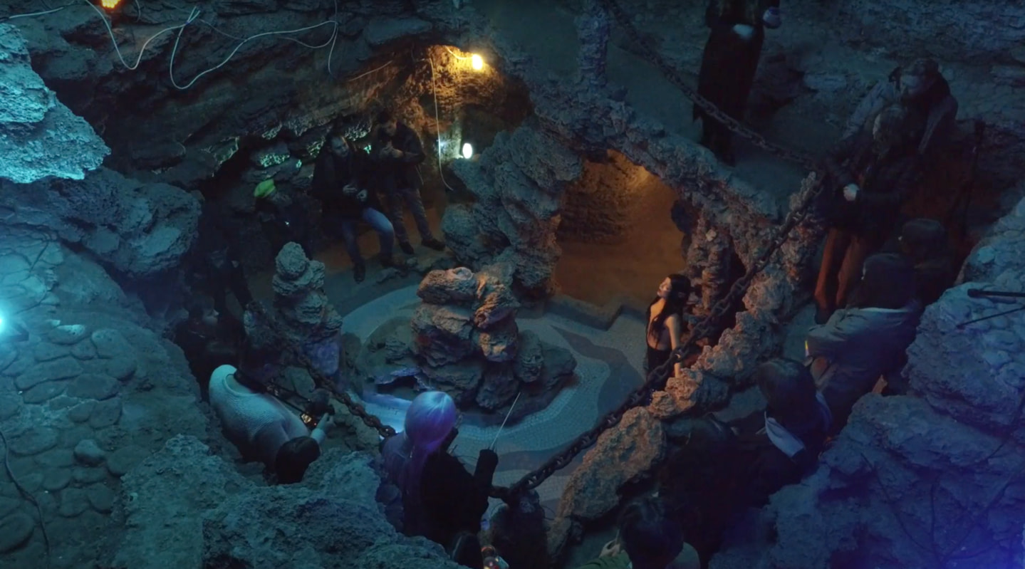 The audience stands along the stone walls of an ancient underground bathhouse, looking down into the center where Sirena’s performance takes place. The space is lit with soft blue and amber lights, creating an atmosphere of ancient ritual and intense listening.