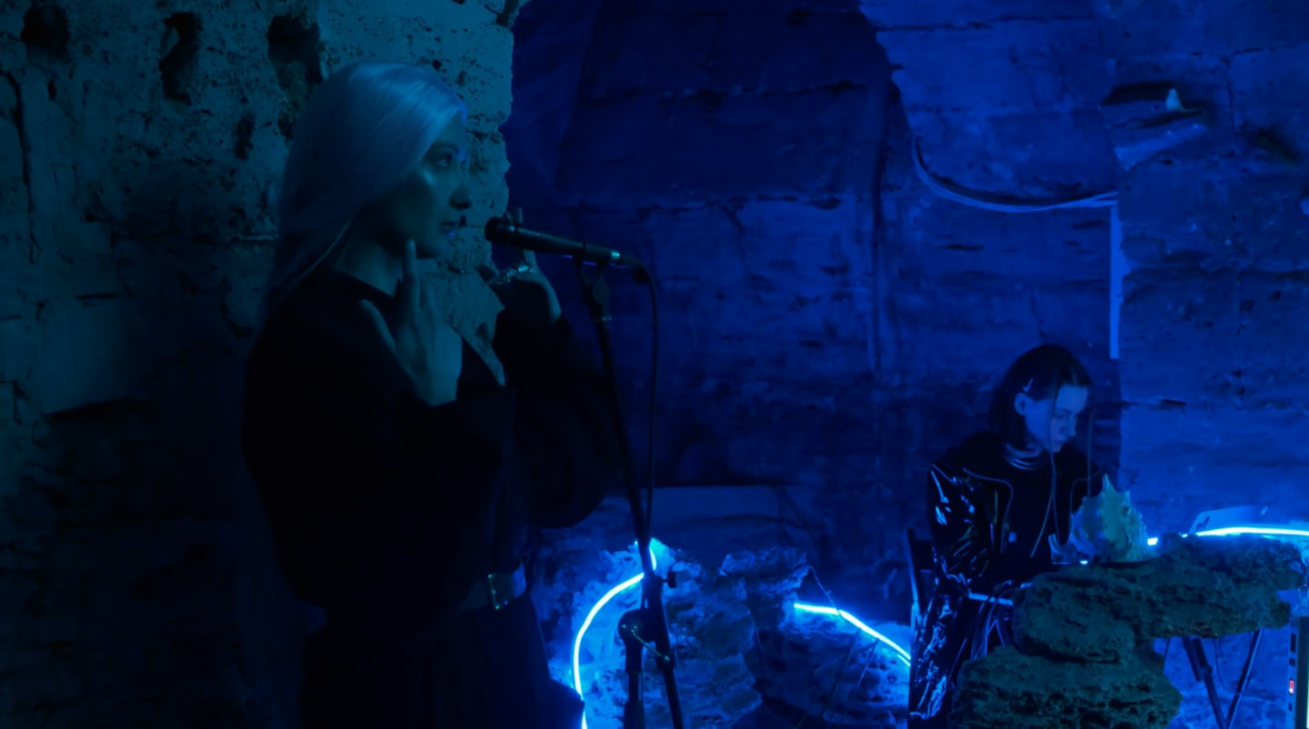 In the blue light of the underground space, an artist with a microphone in the image of an ancient creature, while another participant controls the sound environment with an electronic installation in the performance Sirens