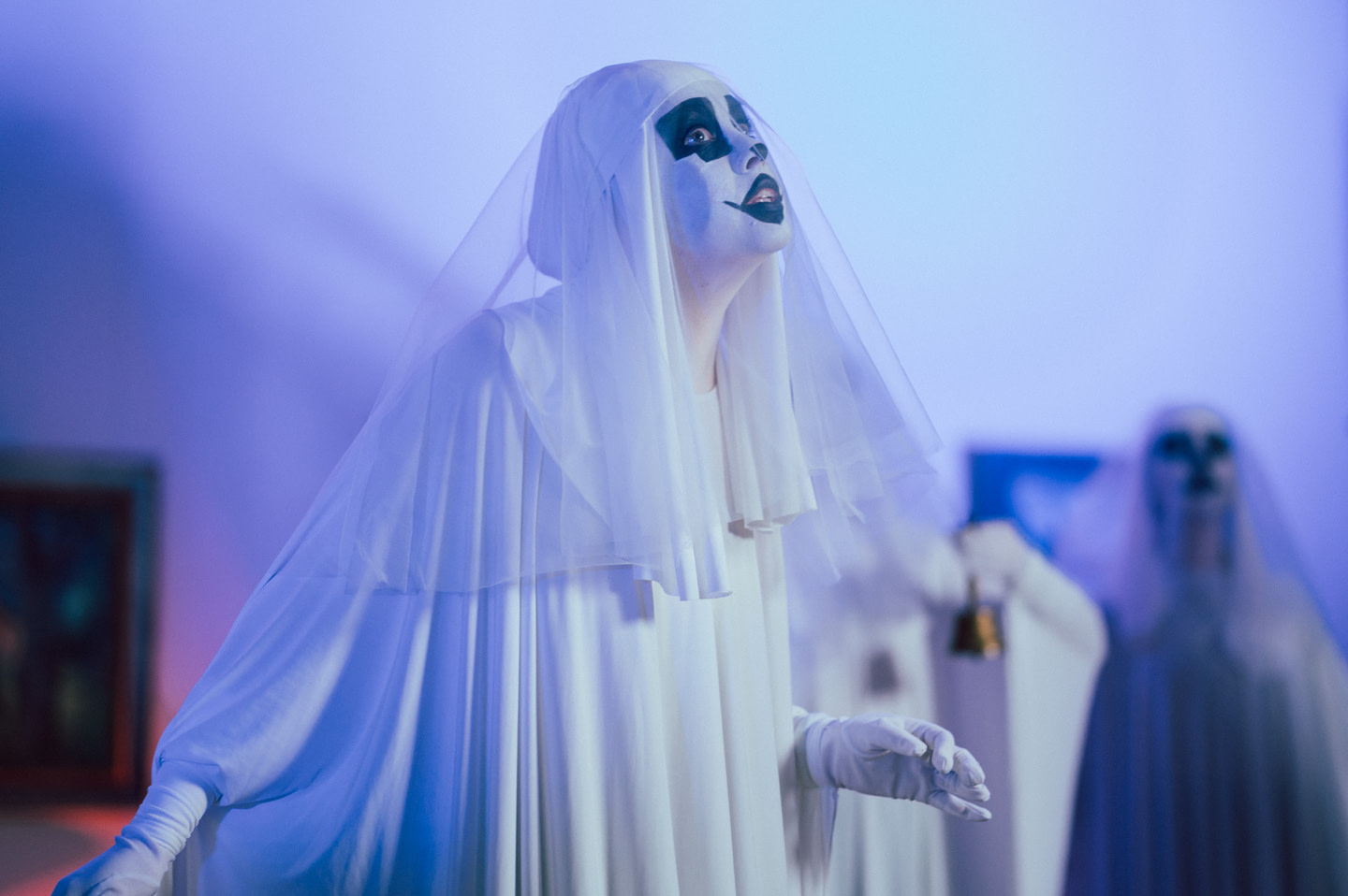 In performance Wolf Man performer in a white outfit with a veil and white gloves, her face painted with black and white makeup. In the background, another figure in a similar costume is visible, holding a bell. The stage is lit with cold blue-violet light, creating a mystical and theatrical atmosphere.
