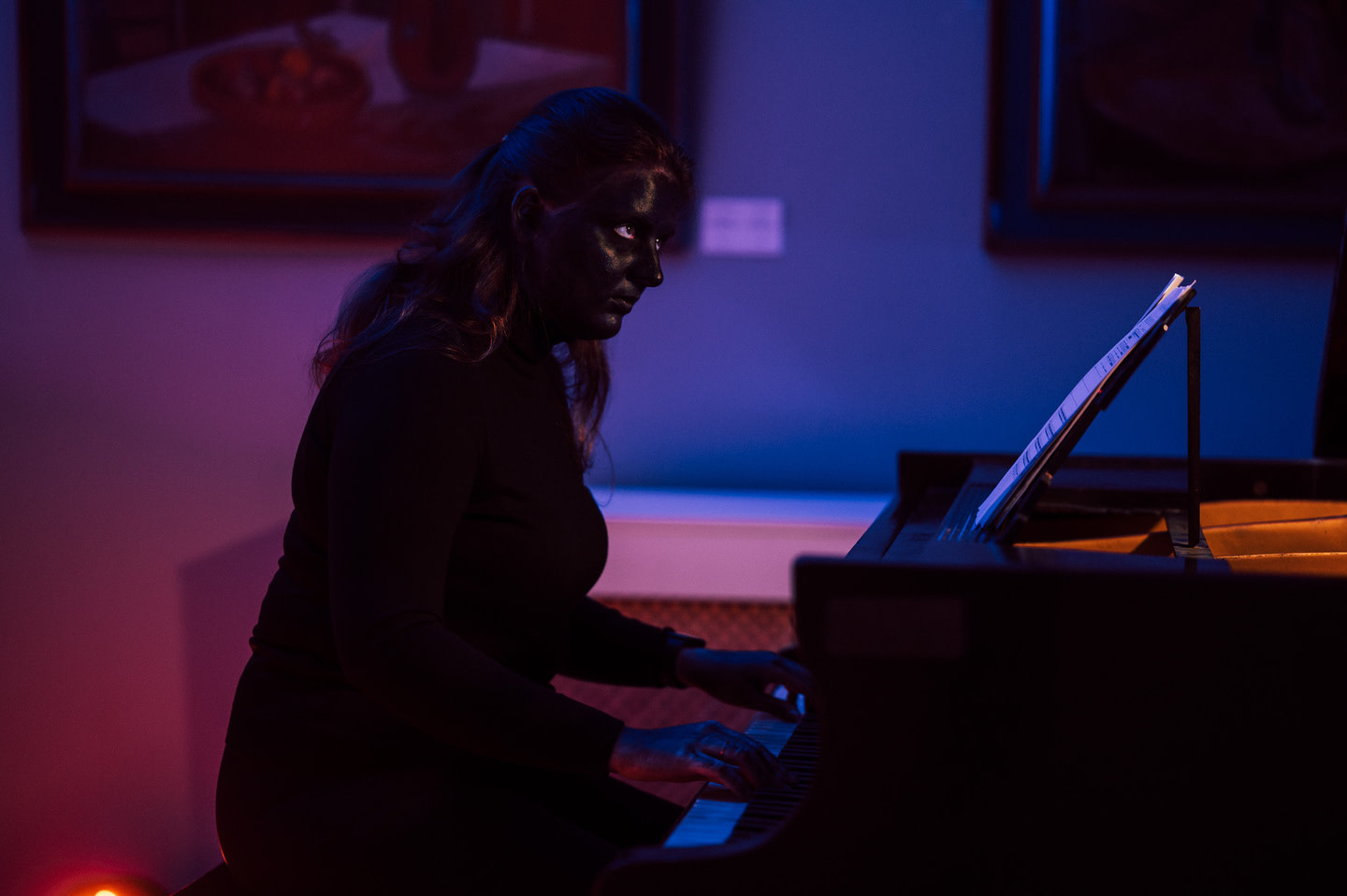 A moment from Malinić's performance "Wolf Man": a woman with her face completely painted in black sits at a piano in dark clothing. She plays the instrument in a deep voice, leaning over the keys.