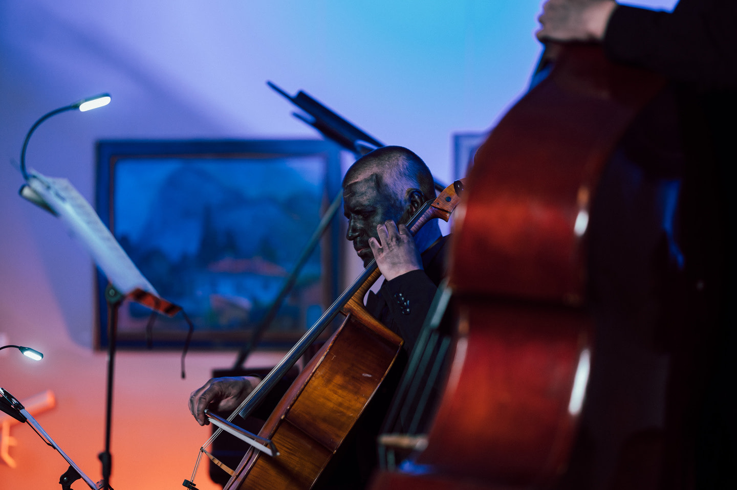 A scene from Malinich's performance "Wolf Man": a musician with a blackened face plays the cello in a semi-dark space flooded with blue-violet light. He is focused on playing, holding the instrument. Framed paintings and music notes on a music stand are visible in the background, and on the right is the part of another musician's double bass.
