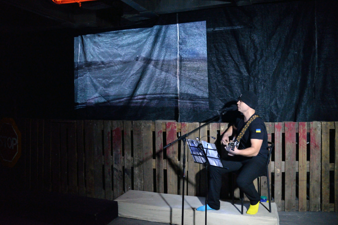 In the performance War Diary, a musician sits on a stage decorated with a wooden fence and black cloth, dressed in black with a Ukrainian flag on his sleeve. He plays the guitar and sings into a microphone, reading from a music stand. He wears multi-colored socks, one blue, the other yellow. Behind him, an image resembling a landscape is projected on the background. A 'STOP' road sign is visible on the left.
