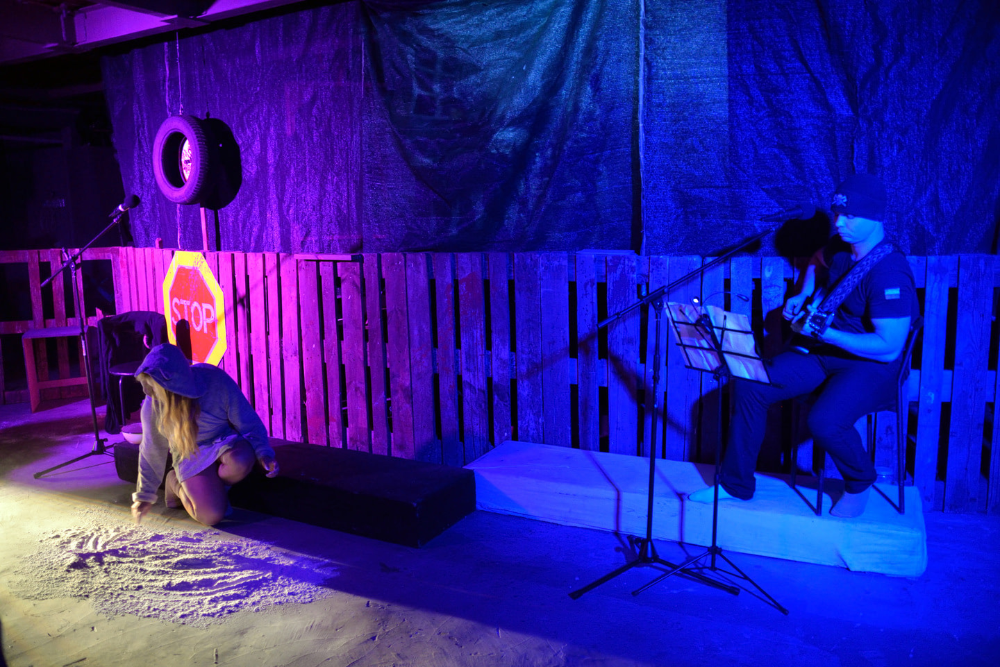 The stage in the performance War Diary is lit with purple and blue lights. A small Ukrainian flag is visible on his T-shirt. On the left, a woman in a gray hoodie and skirt is kneeling on the floor, drawing something in the sand. Behind her is a 'STOP' road sign and a suspended car tire.