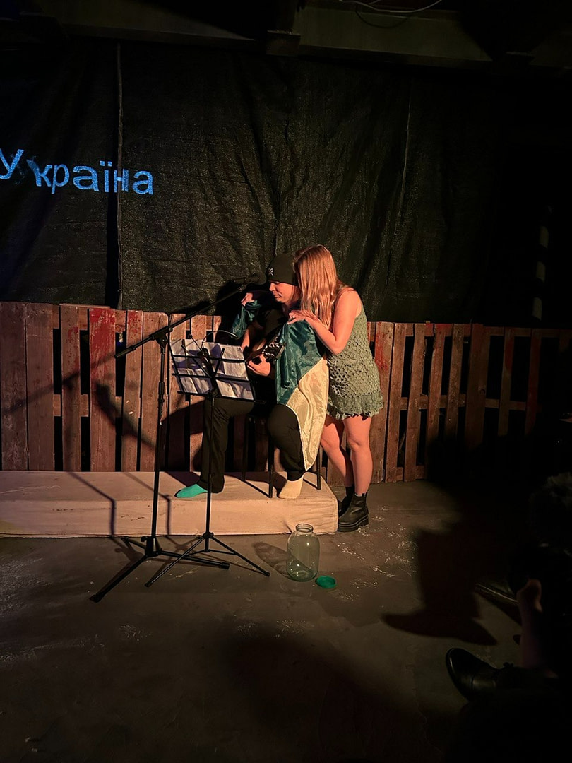 On a dark stage, performance War Diary illuminated by warm light, sits a musician with a guitar, wrapped in the flag of Ukraine. He plays the guitar, reading notes from a music stand. Next to him stands a woman in a green dress, who gently leans towards him, as if singing along or whispering something. Near the stage stands a large empty can with an open lid. On a black background behind, the inscription 'Ukraine' is visible