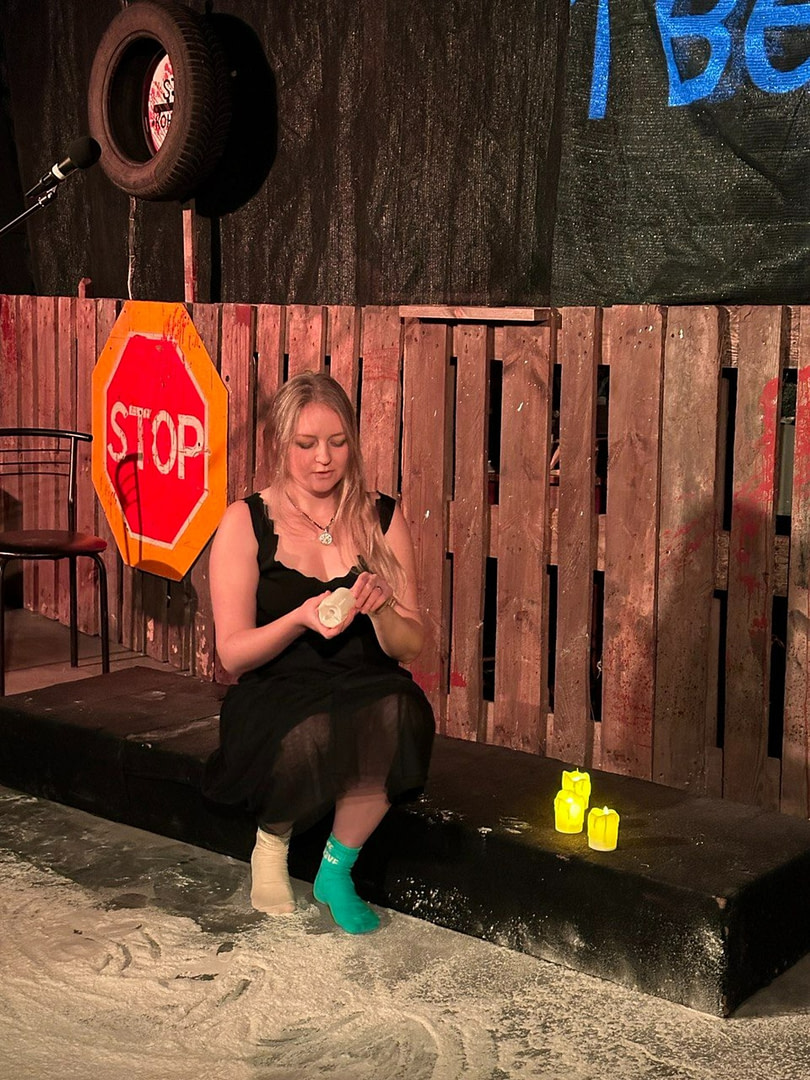 The performer of War Diary sits on stage in a black translucent dress. She holds a rolled up white towel in her hands. She is wearing two different socks - one beige, the other turquoise with the inscription 'LOVE'. Flour is scattered on the ground. and. Behind her is a wooden fence, a 'STOP' road sign, a suspended car wheel and several yellow electric candles glowing near her.